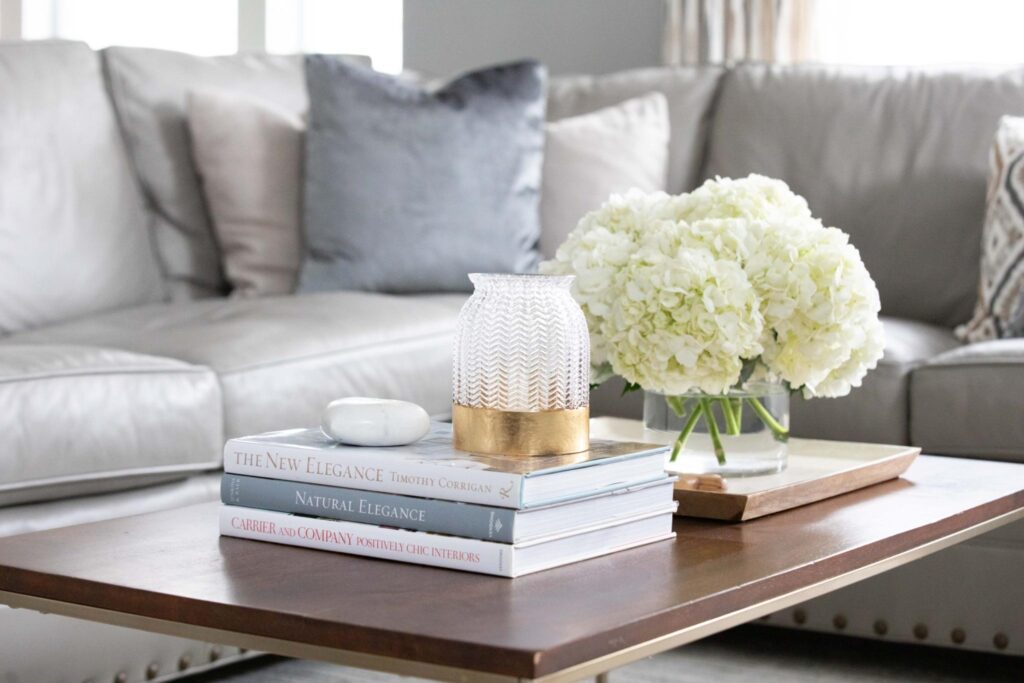A modern luxury living room coffee table styled for a January home refresh. A stack of interior design books sits next to a large clear glass vase filled with lush white hydrangeas and a gold-accented hurricane candle holder, with a plush grey sectional sofa in the background. — JL Signature Designs