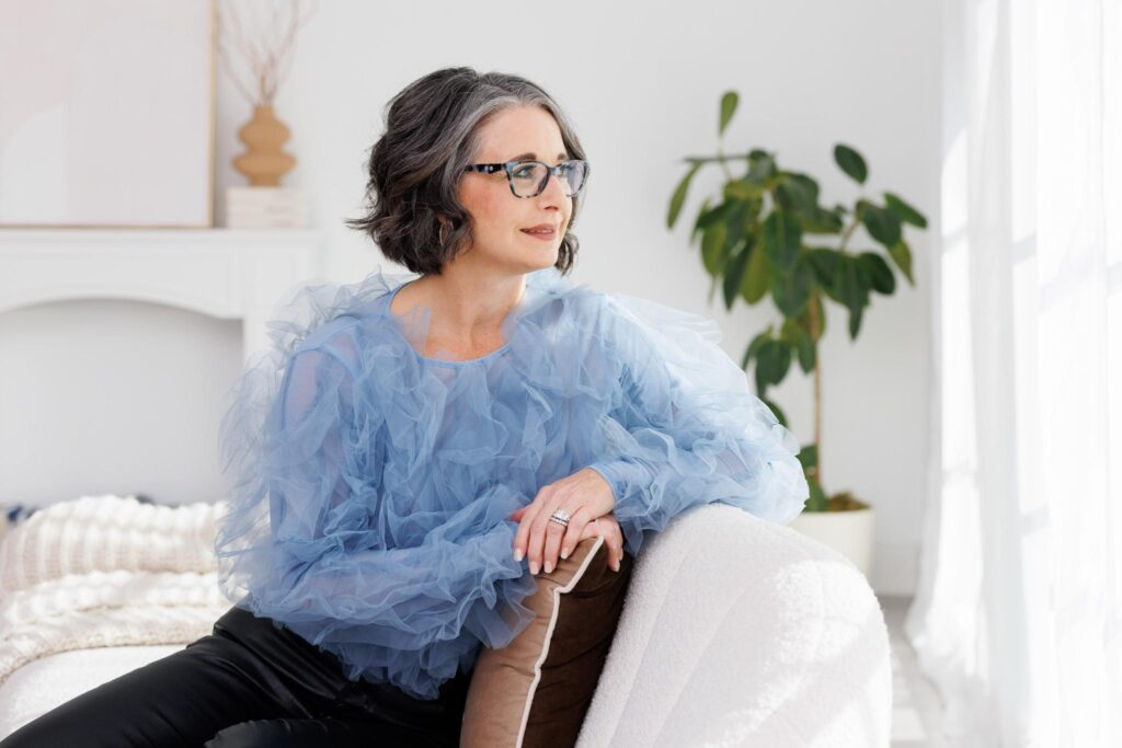 Interior designer Jennifer Looney wearing a sheer blue textured top, smiling and looking out a window while sitting in a modern white chair. — JL Signature Designs