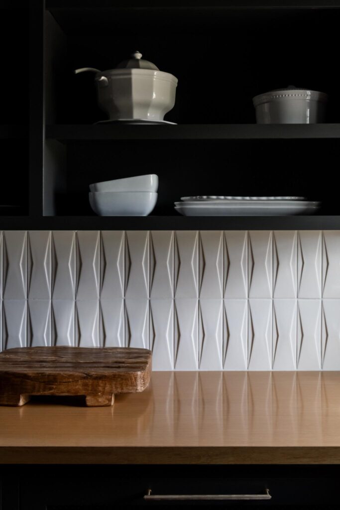 Modern luxury kitchen styling featuring dark cabinetry shelves styled with white ceramic dishware, positioned above a geometric, three-dimensional textured white tile backsplash for stylish design tips. — JL Signature Designs