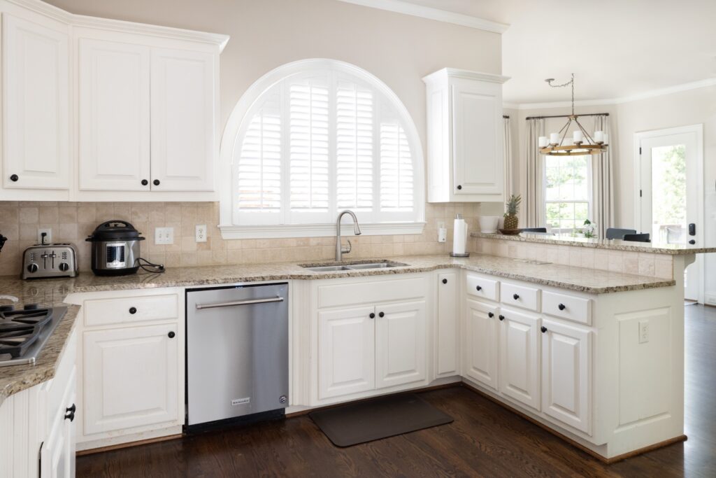 Bright open-concept kitchen before the remodel with high ceilings, featuring a central marble-top counter, integrated stainless steel appliances, and large windows allowing natural light.