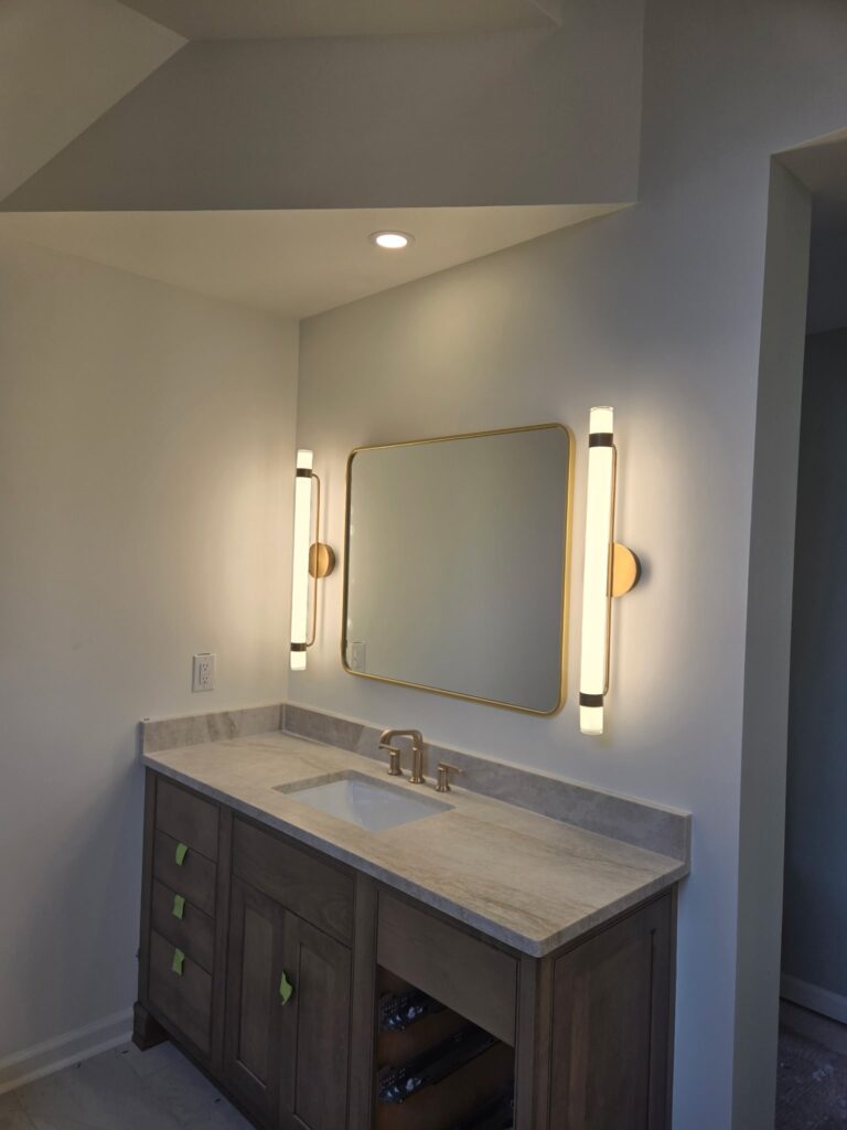 A modern bathroom vanity featuring a natural wood cabinet, a light-colored stone countertop with an under-mount sink, and a large gold-framed rectangular mirror flanked by two vertical gold sconces.