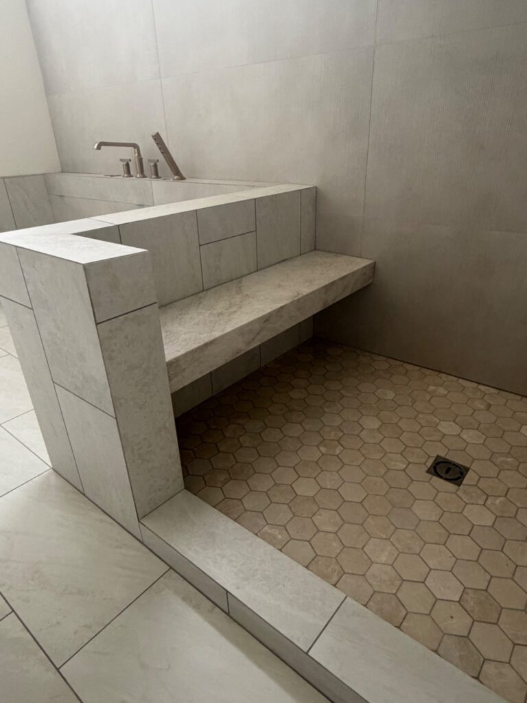 A walk-in shower and bathtub area featuring large-format light gray wall tiles, a built-in shower bench with a marble-look slab, and small tan hexagonal floor tiles.
