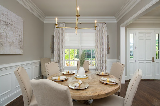 An elegant dining room with a large wooden table, upholstered chairs, a modern statement chandelier, and large windows overlooking lush greenery outside.