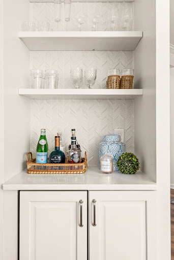 Built-in dry bar featuring white floating shelves with glassware, chevron tile backsplash, and a styled countertop.